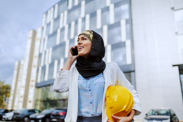 Young positive attractive stylish female muslim architect standing outdoors with helmet under armpit and having business call.