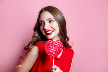Beautiful woman with red lips holding heart shaped lollipop on pink background