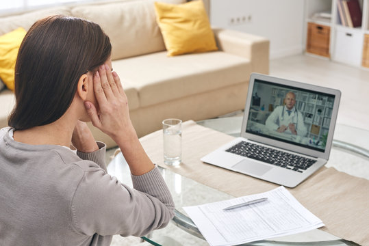 Young sick woman with her hands on temples sitting by table in front of latop during online medical consultation