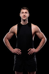 Fototapeta premium Young muscular athlete in black vest and shorts keeping hands on waist while standing in front of camera in isolation