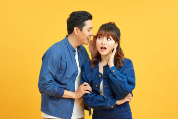 Young man sharing secret with his woman, girl is laughing, yellow studio background