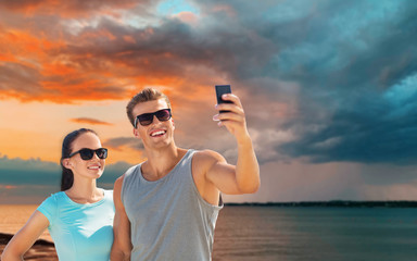 fitness, sport and healthy lifestyle concept - happy couple in sports clothes and sunglasses taking selfie by smartphone over sea and sunset sky background