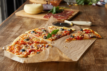 selective focus of delicious italian pizza with olives on baking paper near ingredients and pizza knife on wooden table