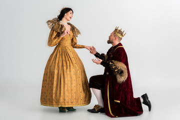 king with crown bending on knee and holding hand of shocked queen on grey background