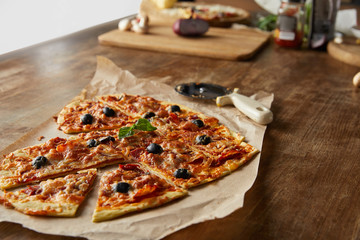 delicious italian pizza in heart shape cut into pieces on baking paper near pizza knife and ingredients on wooden table
