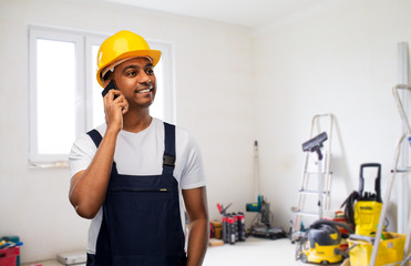 profession, construction and building - happy smiling indian worker or builder in helmet calling on smartphone over room with working equipment at new home or apartment background