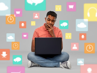 technology and people concept - thinking indian man with laptop computer sitting on floor over mobile app icons on grey background