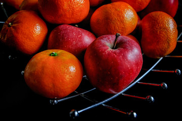 Apples and orange tangerines in a decorative wire basket on a dark background