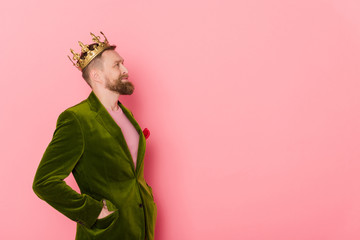 side view of smiling man with crown in velour jacket looking away on pink background
