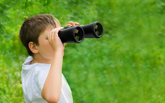 banner size. curious child boy in white t-shirt looking through binoculars in nature green landscape. Explore and discover concept. kid adventure. copy space for text.