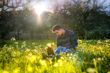 Joven sentado en un campo de flores silvestres, observa algo en el suelo. Detrás de él, el sol...