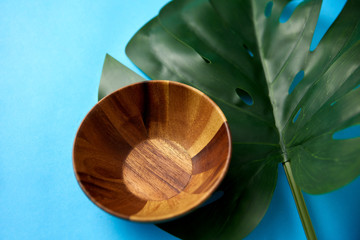 ecology, environmental and exotic concept - close up of wooden plate or bowl and green monstera deliciosa or swiss cheese plant leaf on blue background