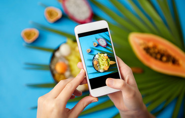 food, technology and healthy eating concept - hands with smartphone taking photo of different exotic fruits mix in bowl with wooden spoon and green fan palm leaf on blue background