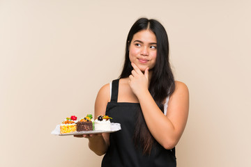 Young teenager Asian girl holding lots of different mini cakes over isolated background thinking an idea