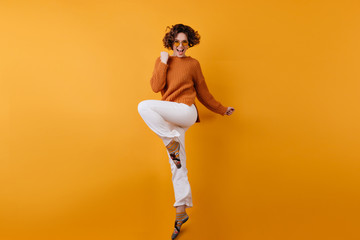 Full-length photo of elegant european female model dancing with excitement. Studio shot of slim brunette girl in white pants jumping with happy face expression.