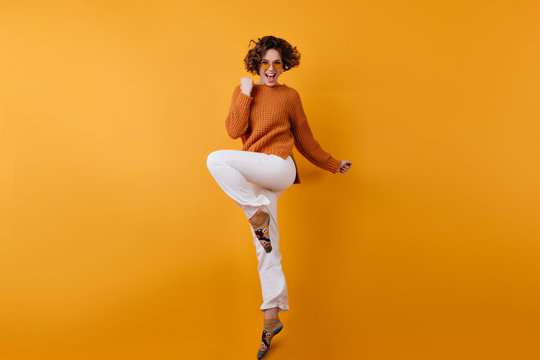 Full-length photo of elegant european female model dancing with excitement. Studio shot of slim brunette girl in white pants jumping with happy face expression.