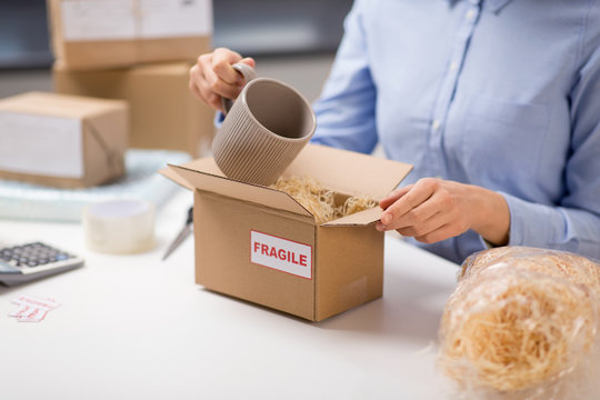 Delivery, Mail Service, People And Shipment Concept - Close Up Of Woman Packing Mug To Parcel Box With Straw Filler At Post Office