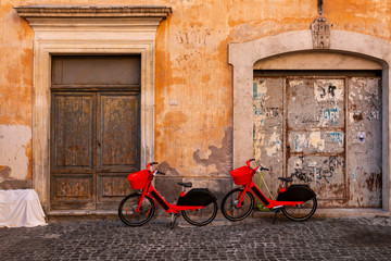 Fototapeta na wymiar Red pedal-assist bikes parked by an old wall. Electric Bicycle sharing system rentals with basket on a sidewalk in street of Rome.