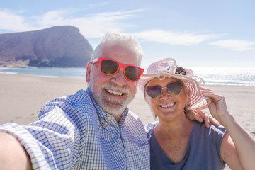 couple of seniors travelling and taking a selfie in a beautiful beach with a mountain at the background - smiling and ejoying looking at the camera wearing sun glasses - vacations lifestyle