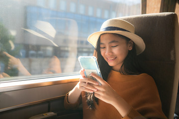 Young asian woman traveler using smartphone in train. Girl traveler smiling holding mobile phone texting online sitting in chair on train in quiet place naer window with warm sunlight..