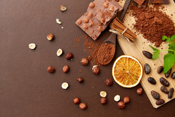 sweets, confectionery and culinary concept - chocolate with hazelnuts, cocoa beans, and powder with dry orange and cinnamon on wooden board with spoon on brown background