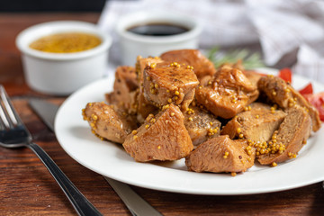 Fried and stewed chicken breast in soy-honey sauce. In the background mustard seeds. On a plate are slices of tomato and razmarin. On a wooden background.