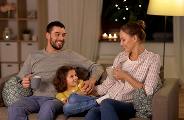 family and people concept - portrait of happy smiling father, mother drinking tea and little daughter sitting on sofa at home at night