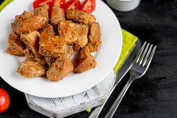 Fried and stewed chicken breast in soy-honey sauce. In the background mustard seeds, a box of vegetables for salad. On a plate are slices of tomato and razmarin. On a black wooden background.