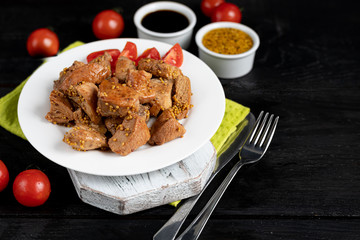 Fried and stewed chicken breast in soy-honey sauce. In the background mustard seeds, a box of vegetables for salad. On a plate are slices of tomato and razmarin. On a black wooden background.