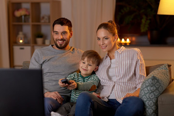 family, leisure and people concept - happy smiling father, mother and little son with remote control watching tv at home at night