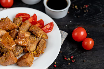 Fried and stewed chicken breast in soy-honey sauce. In the background mustard seeds, a box of vegetables for salad. On a plate are slices of tomato and razmarin. On a black wooden background.