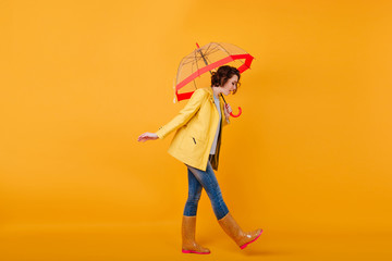 Trendy girl in rubber shoes and yellow jacket looking down while posing with umbrella. Studio shot of curly short-haired woman in jeans walking with parasol. © Look!