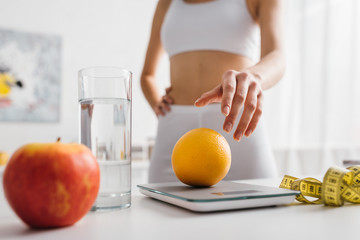 Selective focus of fit sportswoman putting orange on scales near water glass and measuring tape on...