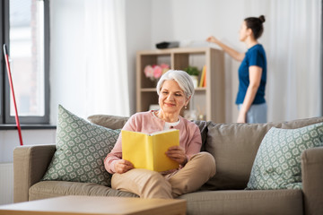 leisure, old age and people concept - happy senior woman reading book and housekeeper cleaning at...