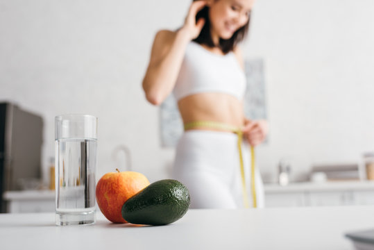Selective focus of glass of water and fruits on table and smiling sportswoman measuring waist with tape in kitchen