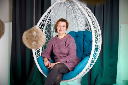 Ugly aged woman in a sweater in a white wicker hanging chair in the room with curtains