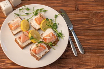Sandwiches with salmon on dark bread, spread with cheese sauce. The meat is wrapped in a slice of daikon and fixed with a decorative clothespin. On a wooden background.