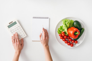 Top view of woman using calculator near notebook and fresh vegetables on plate on white background