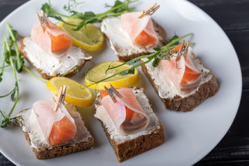Sandwiches with salmon on dark bread, spread with cheese sauce. The meat is wrapped in a slice of daikon and fixed with a decorative clothespin. On a wooden background.
