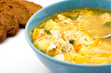 Chicken soup with vegetables in a blue plate on a white table, near a spoon and bread, closeup