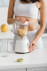 Cropped view of fit sportswoman preparing smoothie with fresh fruits near measuring tape on kitchen table