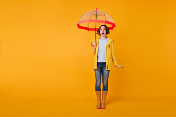 Full-length photo of shocked girl with mouth open standing with umbrella. Trendy young lady in blue jeans posing with amazed face expression in studio.