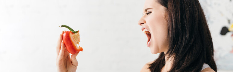 Panoramic shot of young woman grimacing while holding piece of bell pepper in kitchen