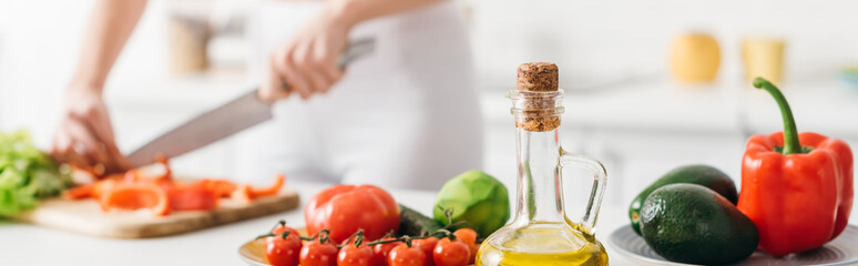 Selective focus of olive oil and fresh vegetables near woman cooking salad on kitchen table,...