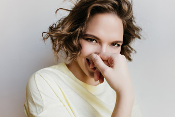 Close-up photo of charming curly lady covering face and laughing. Indoor portrait of carefree girl posing with shy smile on light background.