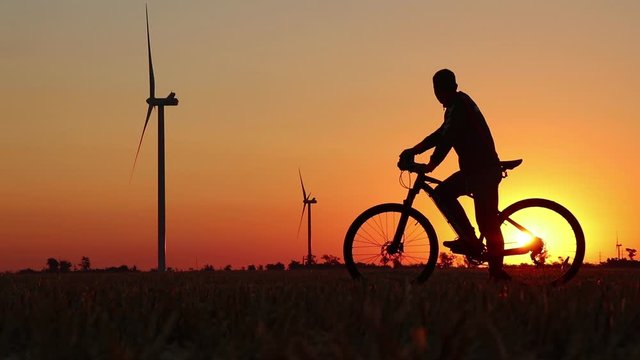 A cyclist stands in a field during the sunset against the background of an orange sky with the sun and windmills