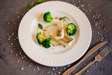 Delicious dish with white fish and broccoli served on the white plate on the grey background. Restaurant menu.