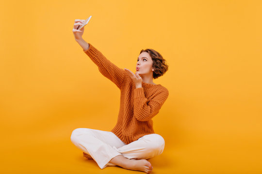 Barefooted young woman in sweater sending air kiss while making selfie. Indoor photo of curly girl in white pants sitting on the floor and taking picture of herself.