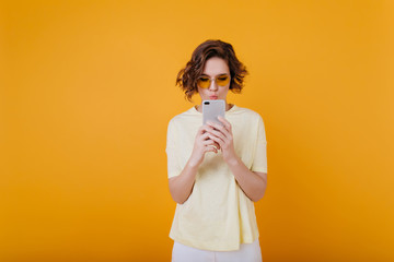 Concentrated curly girl texting message on yellow background. Indoor photo of pretty young woman with wavy hairstyle checking phone mail.