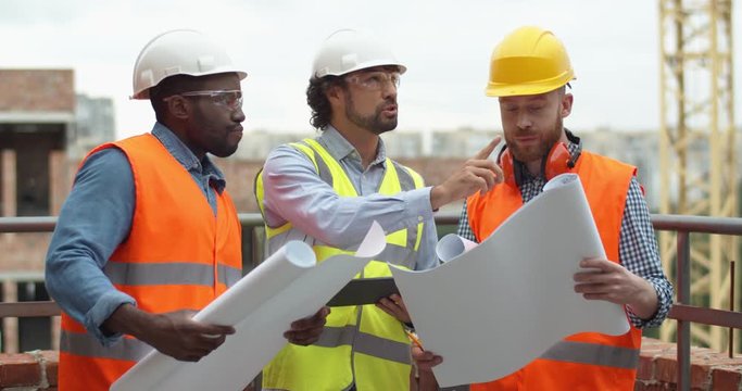 Handsome young foreman in hardhat standing at the industrial construction with two mixed-races workers with drawings in hands and giving them orders what to do.